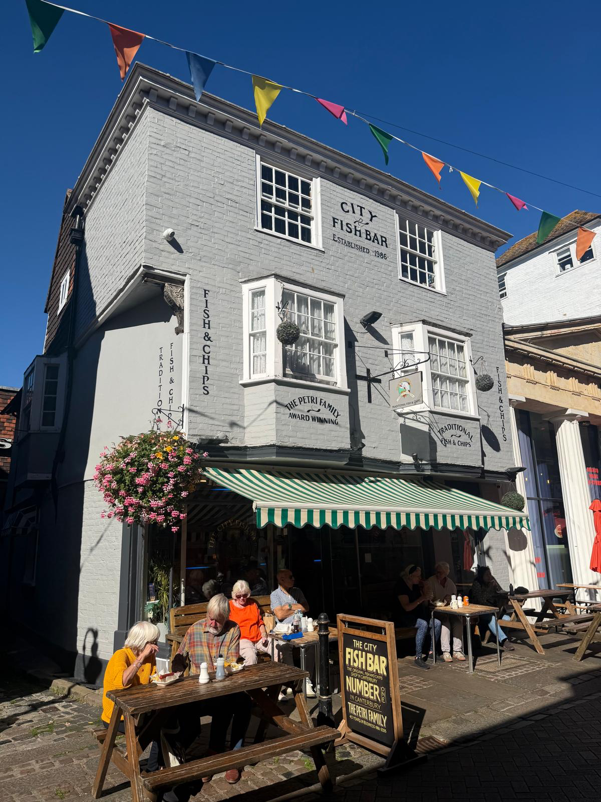 City Fish Bar’s frontage with hanging flowers, striped awning and customers outside